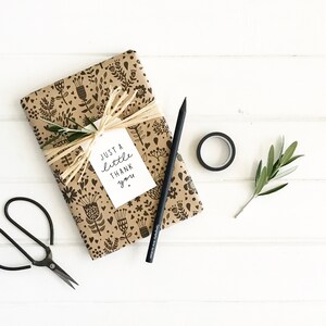 May include: A wrapped gift with a floral pattern, tied with raffia and a tag that reads "Just a Little Thank You." A black pencil, scissors, tape, and a sprig of greenery are arranged on a white wooden surface.