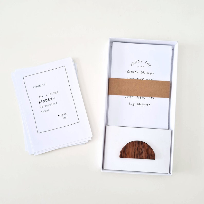 May include: A white box set containing several white cards with black text. One stack of cards reads "Reminder - Talk a little kinder to yourself today. Love me." Another set of cards reads "Enjoy the little things for one day. They were the big things."