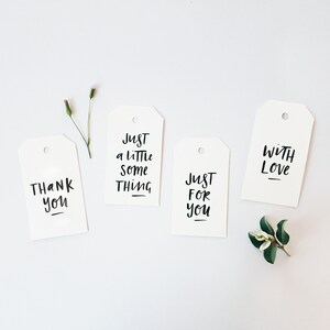 May include: Four white gift tags with black handwritten text: "Thank You", "Just a little something", "Just for you", and "With Love". Small sprigs of greenery and flowers are arranged around the tags on a white background.