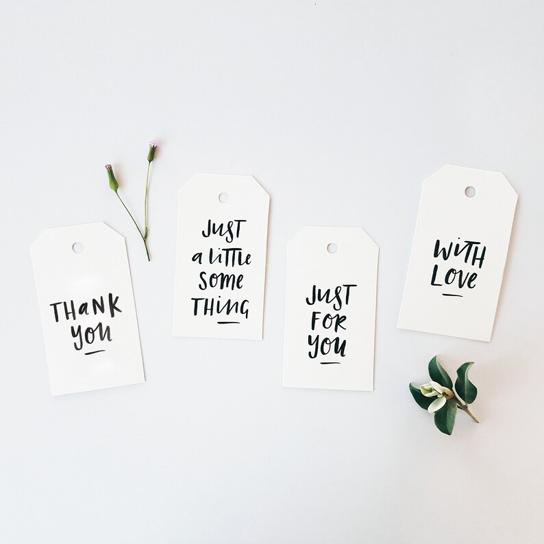 May include: Four white gift tags with black handwritten text: "Thank You", "Just a little something", "Just for you", and "With Love". Small sprigs of greenery and flowers are arranged around the tags on a white background.