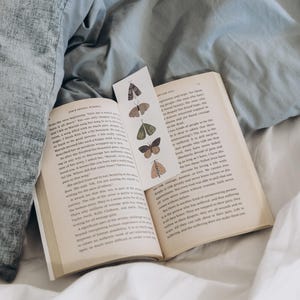 May include: An open book with a bookmark featuring butterfly illustrations. The book is on a bed with a grey duvet and white sheets. The bookmark has various butterfly designs in shades of brown, green, and tan.