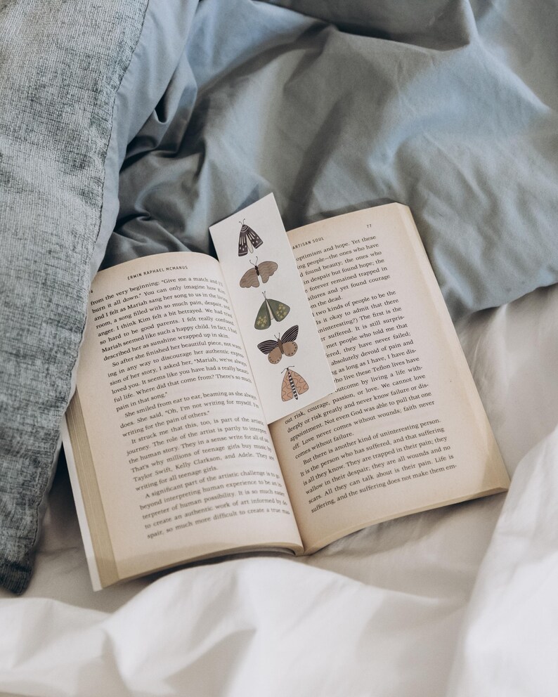 May include: An open book with a bookmark featuring butterfly illustrations. The book is on a bed with a grey duvet and white sheets. The bookmark has various butterfly designs in shades of brown, green, and tan.