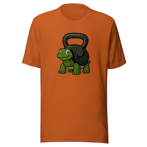 Op de afbeelding: Oranje t-shirt met een cartoon schildpad met een halter. De groene schildpad heeft een zwarte halter op zijn rug. Het ontwerp, gecentreerd op de voorkant, is een speelse kijk op fitness en krachttraining.