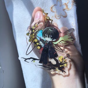 以下が含まれることがあります： 黒髪でダークスーツと赤いネクタイを身に着けたちびキャラが描かれた透明なアクリルキーホルダー。キャラクターは黄色い花が咲く暗い木の枝の上に立ち、金の輪と黄色い翼の要素に囲まれています。