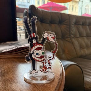Pode incluir: Um expositor de acrílico transparente com uma personagem de desenho animado de smoking e cartola a fazer pino enquanto verte líquido vermelho de um bule para uma chávena. A personagem tem olhos grandes e desiguais e dentes visíveis.