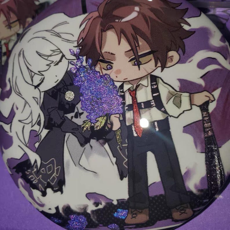 May include: A circular pinback button featuring a stylized anime illustration of a brown-haired character in a white shirt and black harness, standing next to a white-haired figure. The design includes purple glitter accents and a red tie.