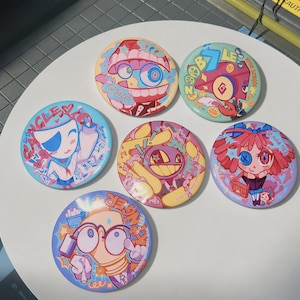 Peut inclure: Six badges ronds à épingle présentant des illustrations de personnages de dessins animés colorés. Les motifs incluent des personnages étiquetés Caine, Zooble, Gangle, Jax, Ragatha et Kinger, chacun avec des palettes de couleurs vives et distinctes.