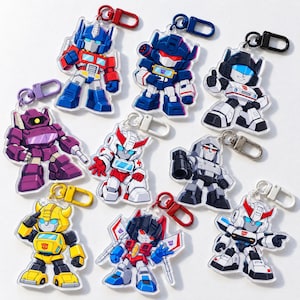 May include: A collection of nine acrylic keychains featuring stylized, chibi-style robot characters from the Transformers franchise. Each keychain includes a colorful plastic or metal lobster clasp and depicts a unique character in a distinct pose.