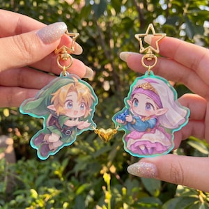 May include: Two acrylic charms featuring anime-style characters, one with blonde hair in a green tunic and the other with a white headpiece and blue and pink dress holding a blue ocarina. Both charms have gold star-shaped clasps and a gold heart connector.