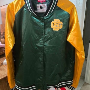 May include: A green and gold satin varsity jacket with a button-up front. The jacket has gold sleeves and a green body with a logo on the chest. The collar and cuffs are striped with black and white.
