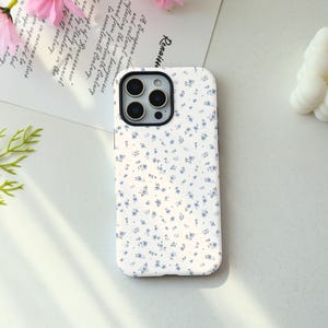 May include: A white smartphone case featuring a repeating pattern of small blue flowers and green leaves. The case has a black camera lens cutout at the top left corner, designed to fit a device with a triple-lens camera system and a flash.