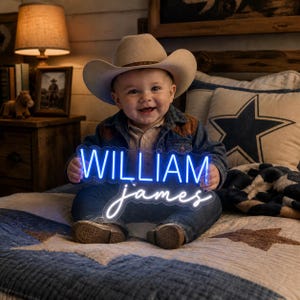 May include: A smiling baby wearing a cowboy hat and denim jacket, holding a blue and white neon sign that reads "WILLIAM james". The baby is sitting on a star-patterned quilt.