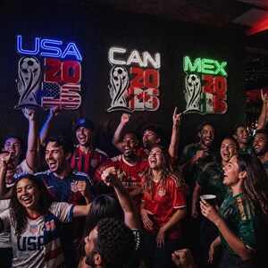 Pode incluir: Um grupo de pessoas celebrando, vestindo camisas de futebol e torcendo. Letreiros de néon acima dizem "USA 2026", "CAN 2026" e "MEX 2026", com gráficos de bolas de futebol e bandeiras. A cena se passa em um bar pouco iluminado.