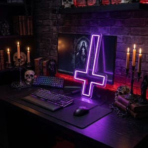 May include: A dark desk setup with a neon purple inverted cross light, a computer monitor, and two keyboards. The desk also features a mouse, mousepad, and lit candles. The background includes a brick wall and bookshelves.