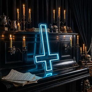May include: A neon blue inverted cross sign is displayed on a black piano. The sign is illuminated with a bright blue light. The piano is surrounded by lit candles in antique candle holders, skulls, and sheet music, creating a gothic aesthetic.