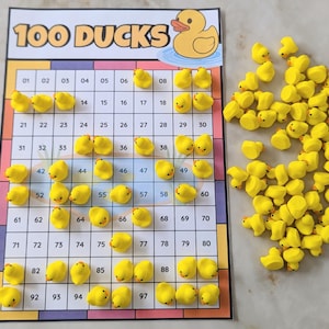 Puede incluir: Un tablero de juego rectangular titulado 100 DUCKS con una cuadrícula numerada del 1 al 100, con una ilustración de un pato de goma amarillo en la parte superior. Numerosas figuras pequeñas de patos amarillos están sobre la cuadrícula y en un montón.