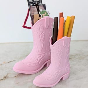 Puede incluir: Dos portalápices con forma de bota vaquera en color rosa claro con patrones de costura estilo western en relieve. La bota más alta contiene una etiqueta negra con el texto THE WORLD y una pegatina de caimán, la más corta contiene varios bolígrafos.