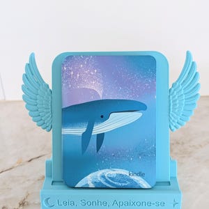 May include: A light blue plastic stand featuring decorative wings on each side and the text "Leia, Sonhe, Apaixone-se" on the front base. The stand holds a Kindle e-reader displaying a graphic of a blue whale swimming through a starry night sky.