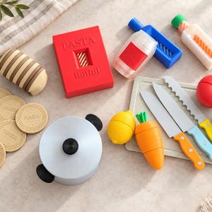 May include: A set of toy kitchen accessories including a red box labeled PASTA Rotini, a silver pot with a black lid, several round tan cookies marked MARIA, a blue grater, a white bottle, three plastic knives, and toy fruits and vegetables.