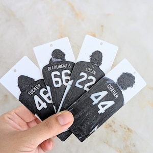 May include: Four rectangular white tags featuring black textured jersey-shaped cutouts. Each jersey displays a name and number: Tucker 46, Di Laurentis 66, Logan 22, and Graham 44. The tags have a small hole at the top for hanging or attachment purposes.