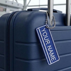 Op de afbeelding: Een marineblauwe harde koffer met een bijpassend rechthoekig bagagelabel bevestigd aan een zilveren kogeltjesketting. Het label heeft een witte rand en de tekst YOUR NAME in witte hoofdletters op een marineblauwe achtergrond.