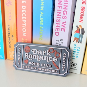 Może przedstawiać: Prostokątna szara tabliczka w kształcie biletu z napisem Dark Romance Book Club Lifetime Membership, Admit One oraz 02631224. Wzór zawiera małą czerwoną czaszkę, czerwone serce i czerwone kajdanki, umieszczone na tle kolorowych książek.