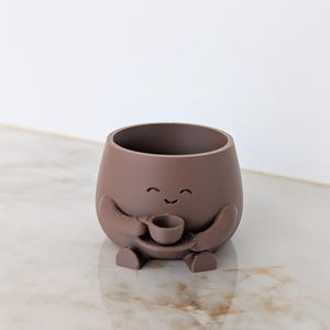 Peut inclure: Un pot de fleurs arrondi en brun mat représentant un personnage sculpté avec un visage souriant. Le personnage a deux bras tenant une petite tasse et une soucoupe assorties devant son corps, avec deux petits pieds visibles à la base du pot.
