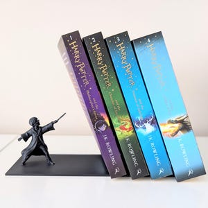 Puede incluir: Un sujetalibros de metal negro con una figura de un mago sosteniendo una varita, que sostiene cuatro libros de Harry Potter titulados Philosopher's Stone, Chamber of Secrets, Prisoner of Azkaban y Goblet of Fire, escritos por J.K. Rowling.