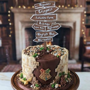 Könnte beinhalten: Eine dekorative Torte mit Harry-Potter-Thema. Die Torte zeigt einen Wegweiser mit verschiedenen Orten, einen schokoladenüberzogenen Abschnitt mit dem Hogwarts-Wappen und dekorative Elemente wie einen goldenen Schnatz und eine Alraune.