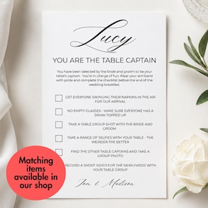 May include: A white rectangular wedding table captain card with black text. The card features the name Lucy, the title YOU ARE THE TABLE CAPTAIN, and a checklist of tasks for guests. A red circular sticker with black text overlays the bottom left corner.