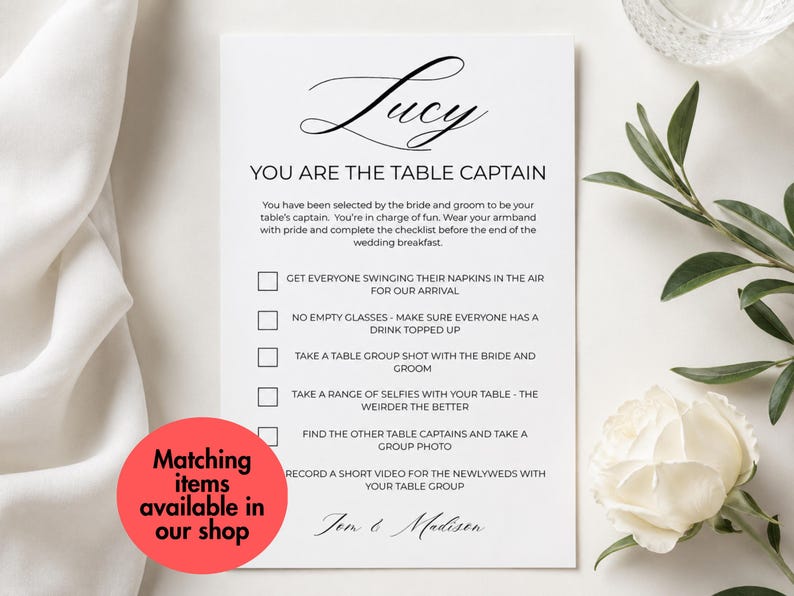May include: A white rectangular wedding table captain card with black text. The card features the name Lucy, the title YOU ARE THE TABLE CAPTAIN, and a checklist of tasks for guests. A red circular sticker with black text overlays the bottom left corner.