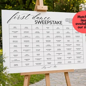 May include: A white rectangular wedding sign on a wooden easel featuring the text first dance SWEEPSTAKE and a grid of song titles and artist names. The sign includes instructions to guess the song and write a name, with the names Lucy and Alex at the bottom.