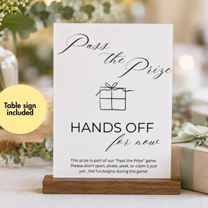 May include: A white rectangular table sign with black text and a line drawing of a gift box. The sign reads Pass the Prize, HANDS OFF for now, and includes instructions for a game. It is held upright by a dark brown wooden base stand.