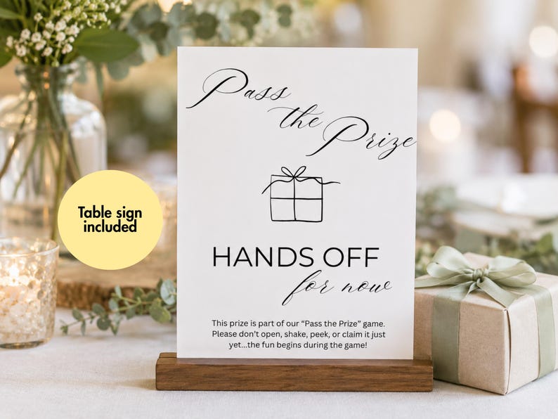 May include: A white rectangular table sign with black text and a line drawing of a gift box. The sign reads Pass the Prize, HANDS OFF for now, and includes instructions for a game. It is held upright by a dark brown wooden base stand.