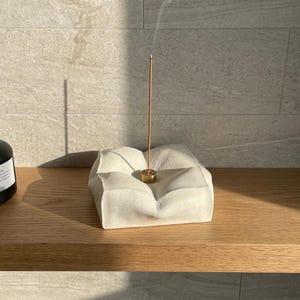 Puede incluir: Un porta incienso blanco texturizado con una base dorada, sosteniendo un palito de incienso encendido. Un difusor de aromaterapia negro está a la izquierda. Los artículos están sobre una estantería de madera contra una pared de azulejos claros.
