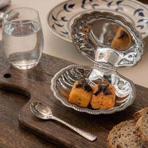 May include: A silver clam shell dish containing two golden-brown food items, a small spoon, and a glass of water on a wooden board. A plate with a blue and white floral design is in the background.
