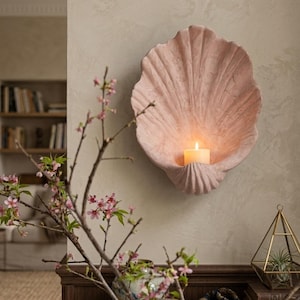 Könnte beinhalten: Eine rosa, muschelförmige Wandleuchte mit einer brennenden Kerze im Inneren. Die Leuchte ist an einer neutralfarbenen Wand angebracht. Ein blühender Zweig und ein geometrisches Terrarium befinden sich im Vordergrund.