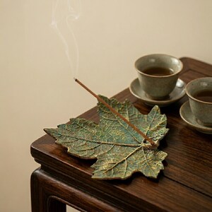 May include: A bronze leaf-shaped incense holder with a lit incense stick, releasing smoke. Two small ceramic teacups and saucers are in the background. The holder rests on a dark wooden table, creating a calm, natural aesthetic.