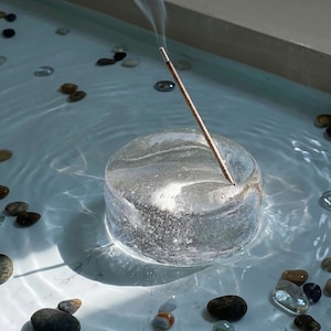 Puede incluir: Un portaincienso redondo y transparente con una varilla de incienso encendida que emite humo. El soporte está en una piscina de agua con pequeñas piedras esparcidas alrededor. El soporte está hecho de un material claro y veteado.