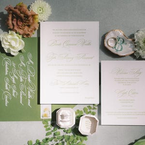 May include: Wedding stationery set featuring pale pink and white cards with gold text. A green envelope with white calligraphy, a ring box, and a seashell ring holder with rings are also present. Floral accents complete the arrangement.
