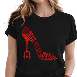 Pode incluir: T-shirt preta com um design de sapato de salto alto vermelho. O texto "THE DEVIL WEARS PRADA" está incorporado no contorno do sapato, com um tridente como salto. A t-shirt tem mangas curtas.