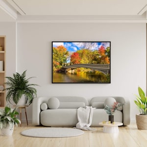 Op de afbeelding: Een ingelijste landschapsprint met een stenen brug over een kalm water, omringd door bomen met levendige herfstkleuren. Het kunstwerk hangt boven een lichtgrijze bank in een moderne woonkamer.