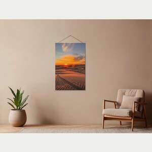 Puede incluir: Una impresión vertical de un paisaje desértico al atardecer. La imagen muestra dunas de arena con huellas de neumáticos, bajo un cielo de tonos naranja, amarillo y azul. La obra de arte está colgada en una pared beige.