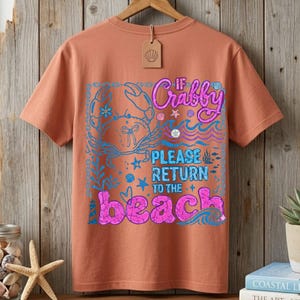 以下が含まれることがあります： コーラル色のTシャツ。ビーチをテーマにしたグラフィックデザイン。青いカニのイラスト、波、ピンクと青で「If Crabby Please Return to the Beach」の文字が描かれています。貝殻のタグが襟からぶら下がっています。