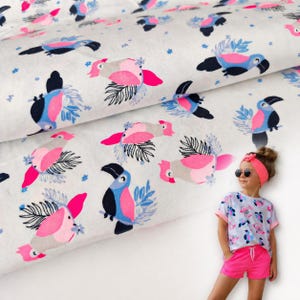 Può includere: Tessuto bianco con un motivo ripetuto di tucani rosa, blu, neri e grigi. I tucani sono circondati da foglie blu e piccoli fiori blu. Una persona giovane indossa una maglietta abbinata e pantaloncini rosa.