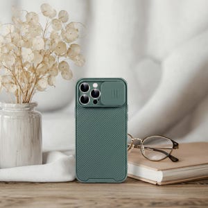 May include: A teal phone case with a camera cover and a textured, diagonal stripe design. The case is next to a pair of gold-rimmed glasses and a closed book. A vase of dried flowers sits in the background.