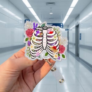 Pode incluir: Um porta-crachá com um design floral de caixa torácica. A caixa torácica é branca com contornos pretos, preenchida com flores coloridas. O porta-crachá tem uma borda brilhante e uma alça de plástico transparente.