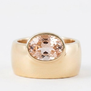 May include: A wide gold band ring featuring a central oval-cut peach-colored gemstone set in a smooth bezel setting. The ring has a polished finish and a thick, solid construction that highlights the soft, warm tones of the faceted center stone.