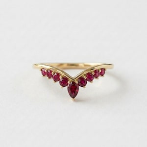 May include: A gold-colored ring with a V-shaped design, adorned with multiple small, round, deep red gemstones and a single, larger, teardrop-shaped red gemstone at the center. The ring is set against a plain white background.