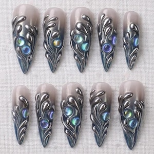 May include: Set of ten press-on nails with a pearlescent base and ornate silver-colored, swirling designs. Each nail features iridescent, jewel-toned accents. The nails are almond-shaped with a pointed tip.